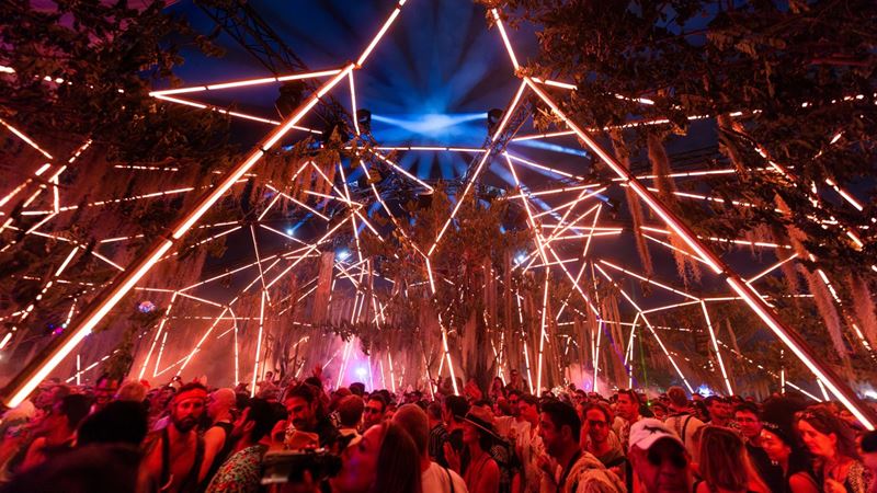 Party inside a dome surrounded by lights and sounds at this year's new Polygon Live LDN. Credit: Marc. Image courtesy of Polygon Productions. People partying under red led lights and trees.
