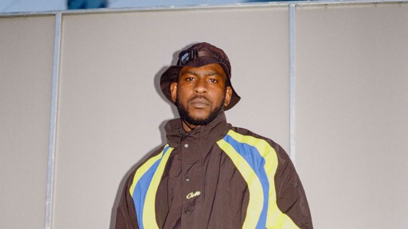 See Skepta and his curated lineup take over Crystal Palace Park at Big Smoke Festival this summer. Credit: Huxley. Image courtesy of Huxley. A photo of Skepta backstage at Big Smoke Festival wearing a bucket hat and Corteiz tracksuit taken on a film camera