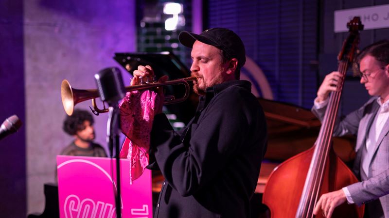 See live jazz bands take to the stage at Soho Jazz Festival this autumn. Credit: Soho Live Studios. Image courtesy of Soho Jazz Festival. A man blowing into a trumpet onstage with other musicians during Soho Jazz Festival