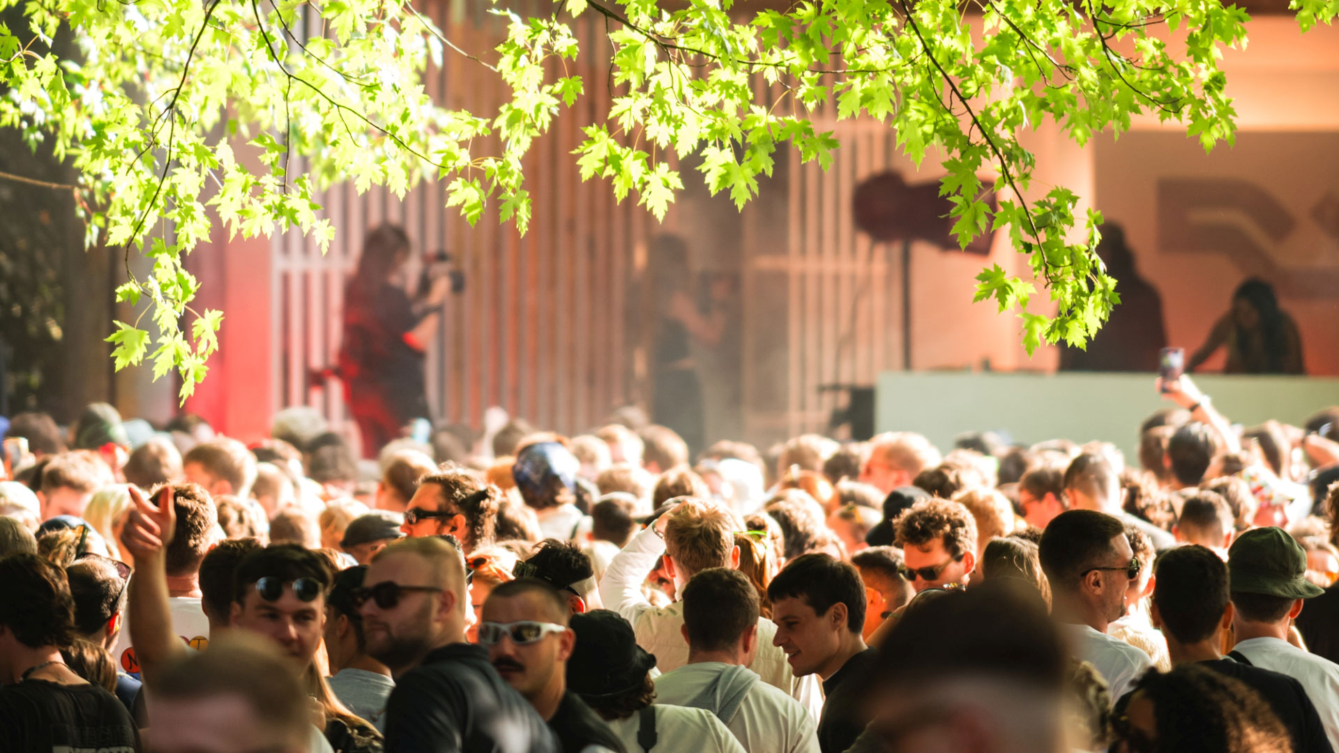 Party at Waterworks in west London's Gunnersbury Park. Image courtesy of Percolate Music. A photo of a crowd of festival-goers in the sunshine at Waterworks festival with two DJs playing onstage in the background and branches of a tree hanging in the top of the frame