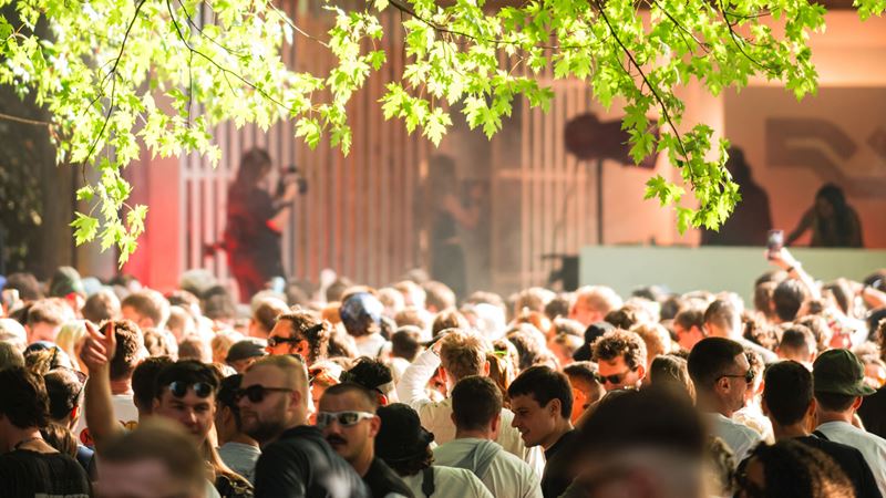 A photo of a crowd of festival-goers in the sunshine at Waterworks festival with two DJs playing onstage in the background and branches of a tree hanging in the top of the frame