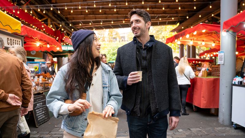 two people walking through a covered food market, with pop up stands decorated in fairy lights