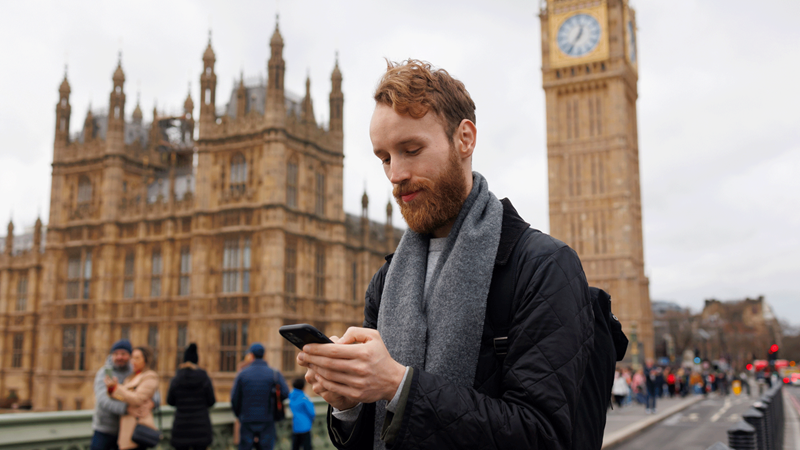A man with black coat and grey scarf looks down at his phone in front of Big Ben