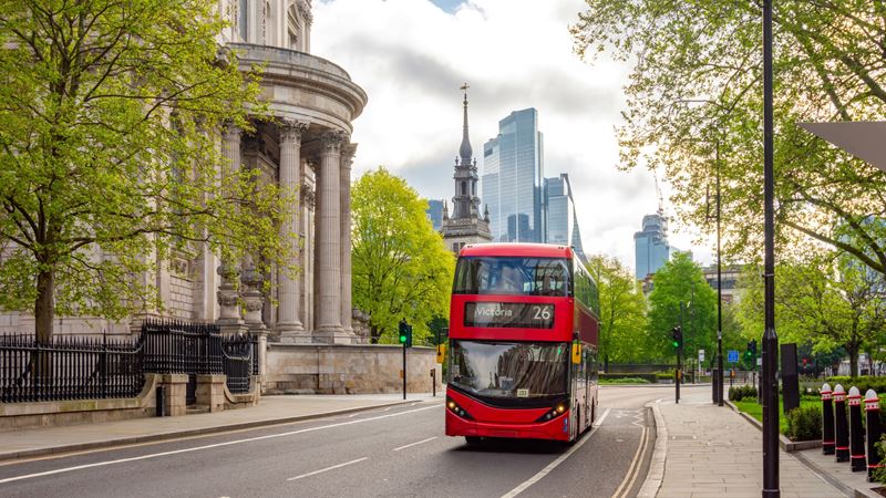 A red double decker bus drives along a road in London with a stately building and modern offices in the background and leafy trees lining the road