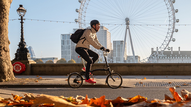 Getting around London on a bicycle. Image courtesy of visitlondon/Antoine Buchet. A man cycling on a bridge with the London Eye in the background and autumn leaves in the foreground.