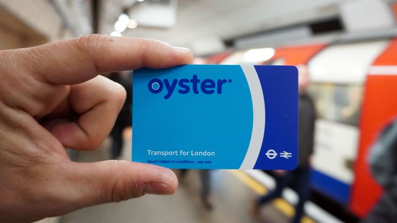 Person holding a blue London transport Oyster card with a London Underground train arriving at a Tube platform in the background.