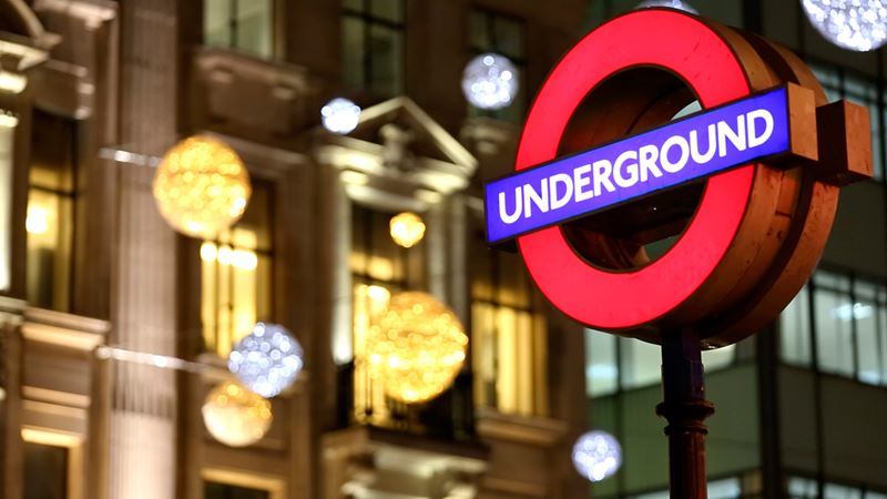 A London Underground sign lit up at night, with Christmas lights shining in the background. 