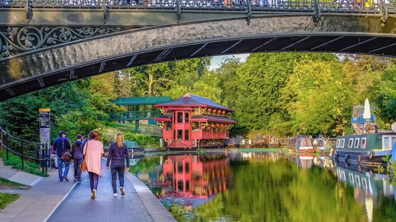 Look out over the picturesque Regent's Canal from the floating Feng Shan Princess. Credit: Shutterstock. Image courtesy of Shutterstock. A photo of the back of two women walking along Regent's Canal with a floating restaurant and houseboats and a bridge in the foreground