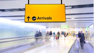 A yellow "Arrivals" sign in an airport, above a travelator walkway in an airport corridor.