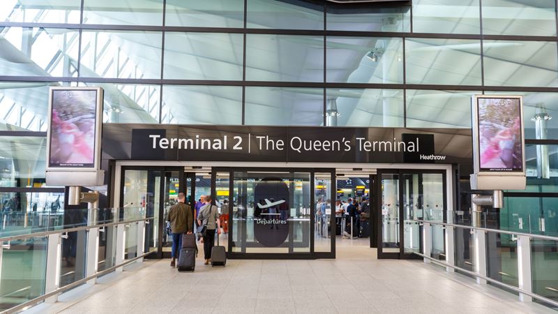 A photo of people wheeling suitcases through the entrance to The Queen's Terminal at Heathrow Airport