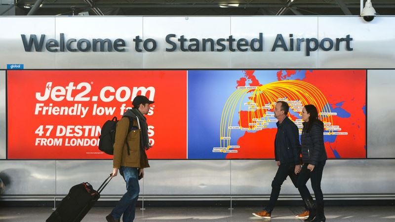 People walk in front of a Welcome to Stansted Airport sign with a digital advert for Jet2 in the background