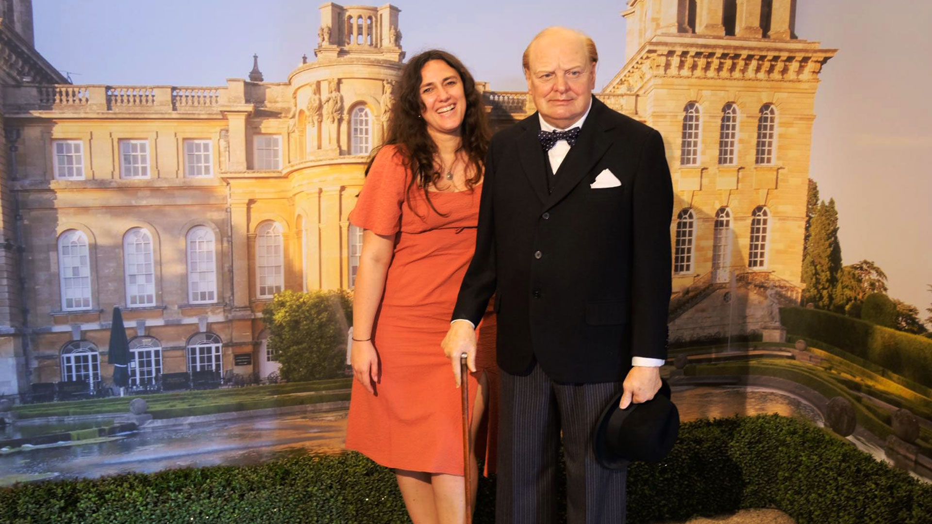 Don’t forget to strike the pose with Winston Churchill at Blenheim Palace. Image courtesy of Carole Rocton. A woman in a rose dress stands next to a wax figure of Winston Churchill with Bleinheim Palace in the background.