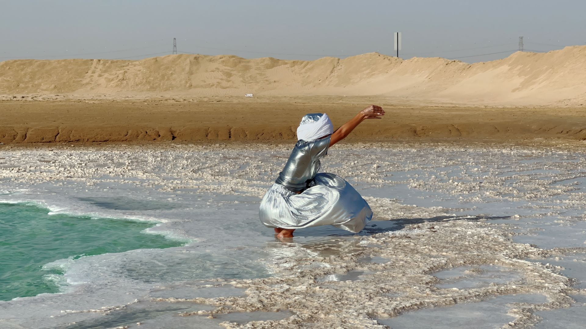 SALT COSMOLOGIES - HylozoicDesires. Soap of the sages, sun of the sea, 2024. Image courtesy of Somerset House. A person stands on the sand by the ocean in an art installation called SALT COSMOLOGIES- HylozoicDesires, Soap of the sages, sun of the sea.