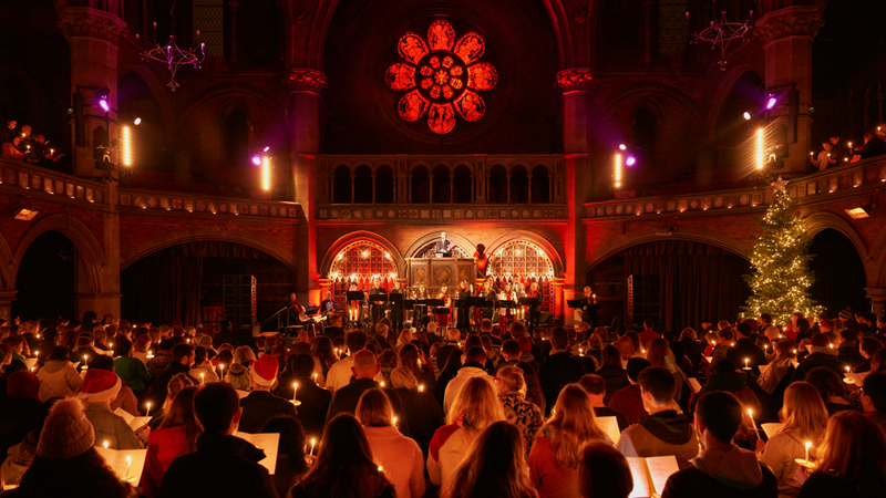 Gather at Union Chapel in Islington for a night of Christmas carols, music, spoken word and performances. Credit: Emile Holba. Image courtesy of Union Chapel. Crowds of people stand on across two levels at Union Chapel as a round red stained glass window illuminates the whole room with performers and musicians stood on the stage