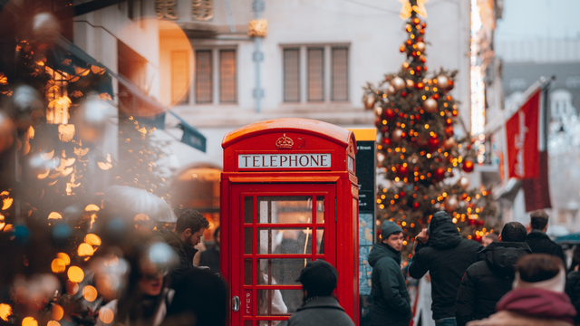 A crowd walks through London past a bright red telephone box and christmas trees.