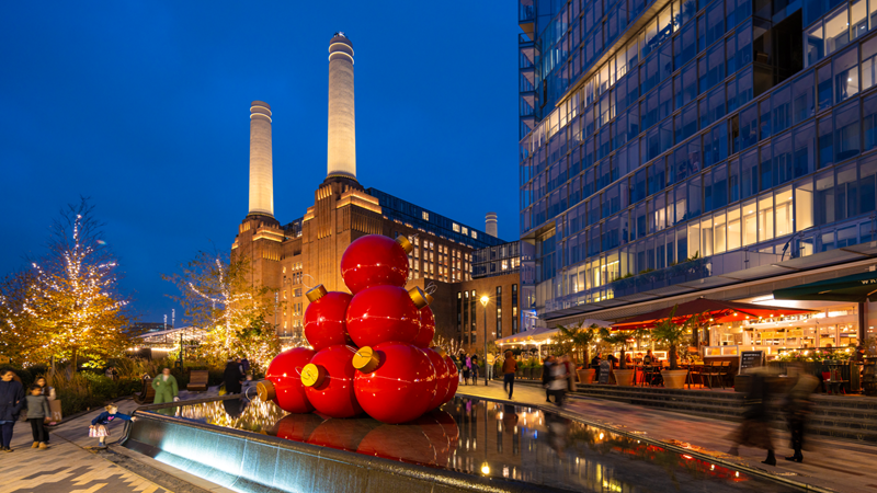 See Battersea Power Station transformed for Christmas this festive season. Credit: Charlie Round-Turner. Image courtesy of Battersea Power Station. A pile of giant red bauble decorations stood in front of Battersea Power Station at blue hour as glowing lights and Christmas trees light up the area