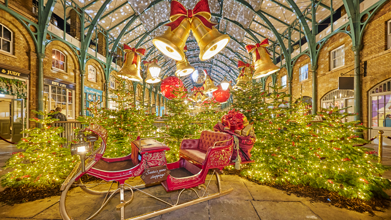 Soak up the season in Covent Garden. Credit: Covent Garden. Image courtesy of Covent Garden. A red sleigh with gold details in the Covent Garden market surrounded by Christmas trees with twinkling lights with giant gold bells with bows hanging above