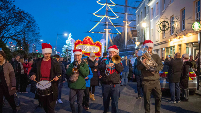 Join the crowds that flock to Greenwich for the annual Light Parade to mark the start of the festive season. Credit: Ed Simmons Photography. Image courtesy of Visit Greenwich/Greenwich Market. A four piece band walk down the street in front of Greenwich wearing Christmas hats surrounded by people holding lanterns and lights, with a view of Cutty Sark in the background decorated with lights in the shape of a Christmas tree