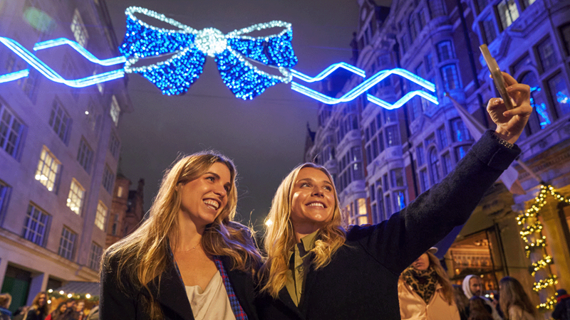 Snap a photo in front of Mayfair Christmas lights. Credit: Simon Jacobs/PA Wire. Image courtesy of Four Agency. Two female friends take a selfie in front of Mayfair Christmas lights, featuring a bright blue bow
