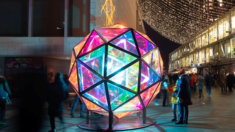 See the South Bank area illuminated this winter with Winter Light and Christmas lights. Credit: South Bank London. Image courtesy of South Bank London. A large round light up installations made up of triangular glass panels emitting pink, blue, green, orange and yellow lights as people stand around it and twinkling string lights glow in the distance