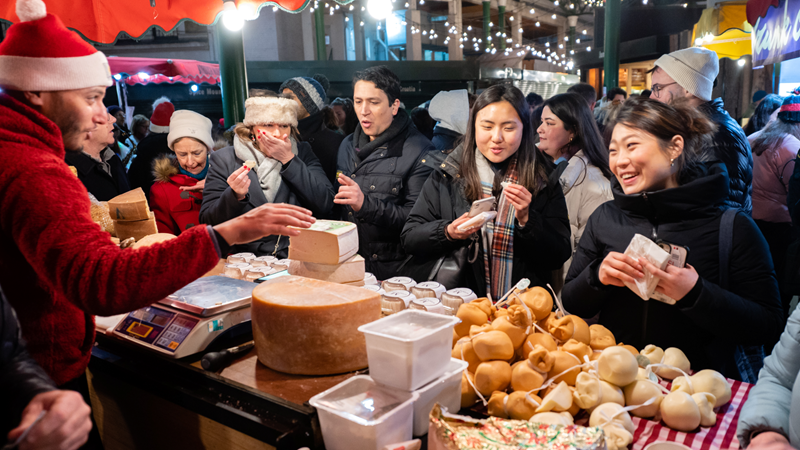 Borough Market is a must-visit destination for foodie lovers at Christmas! Credit: Borough Market. Image courtesy of Borough Market. Crowds of people wrapped in warm clothes queue for a cheese stall at Borough Market stocked with piles of cheeses of different shapes, sizes and colours whilst a man behind a counter wearing a Christmas hat weighs cheese.