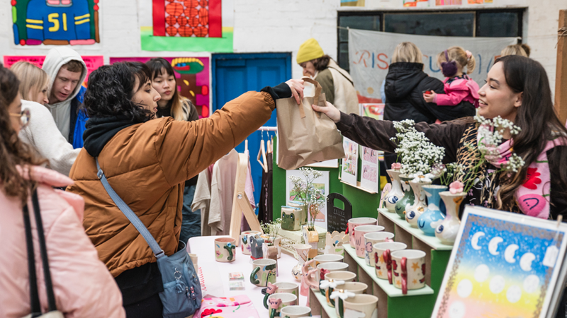 Browse the stalls of emerging artists and independent sellers at the DIY Christmas Art Market in south London. Credit: Jérôme Favre. Image courtesy of DIY Art Market. A young woman in a brown puffer jacket with a blue shoulder bag takes a brown paper bag filled with goodies from an independent seller at a market, with her stall full of goodies like colourful handmade mugs and painted canvases