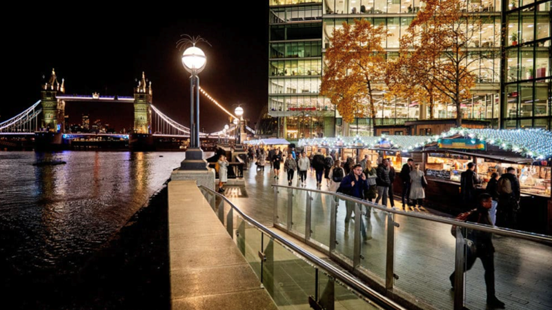 Take in the views of Tower Bridge and the river Thames from Winter by the River Christmas market. Credit: Winter by the River. Image courtesy of Winter by the River. People walk past stalls selling Christmas fare and covered with LED lights next to the river Thames at night with Tower Bridge in the distance