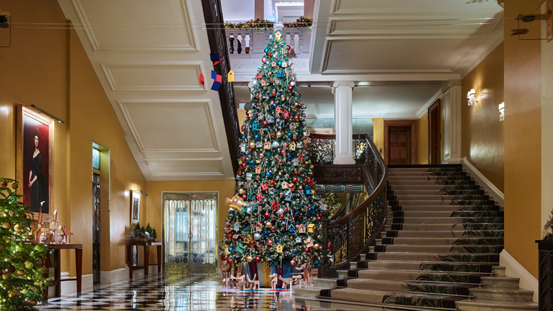 Step inside the Claridge's lobby and you'll be greeted by this year's whimsical Christmas tree designed by Paul Smith. Credit: Claridge's. Image courtesy of Claridge's. The Claridge's hotel lobby dressed up for winter featuring a large curved staircase and tall Christmas tree in the middle of the room, covered in warm twinkling lights and decorations designed by Paul Smith, in the form of birdhouses, clocks, colourful baubles and more.