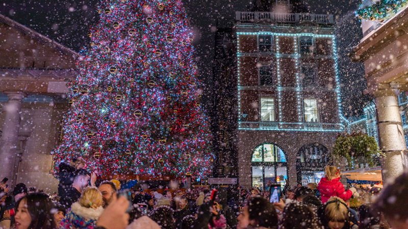 A photo of people dressed for winter at Covent Garden as snow falls with a Christmas tree in the background covered in red and blue twinkling lights