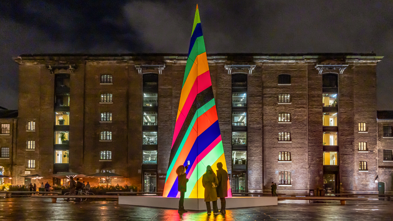 You can't miss the kaleidoscope of colours that are set to lighten up the wintry nights at Granary Square. Credit: John Sturrock. Image courtesy of King's Cross. A large abstract Christmas tree stands in Granary Square on a dark night giving off light in many different vibrant colours as three people stand in front of it