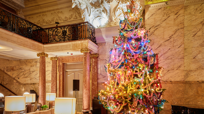 See The London EDITION Christmas tree take centre stage with a good helping of tinsel. Credit: Henry Mills. Image courtesy of The London EDITION. A Christmas tree stands in the lounge of The London EDITION covered in multi-coloured sparkly tinsel and lights. A pile of presents sit below the tree and there's a corner of a piano on one side and sofas on the other, with marble pillars holding up a viewing gallery up above