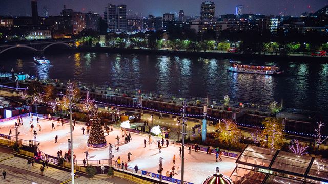 An overview of the Glide Battersea ice rink, illuminated at night and located on the banks of the Thames with a view of the London skyscrapers.