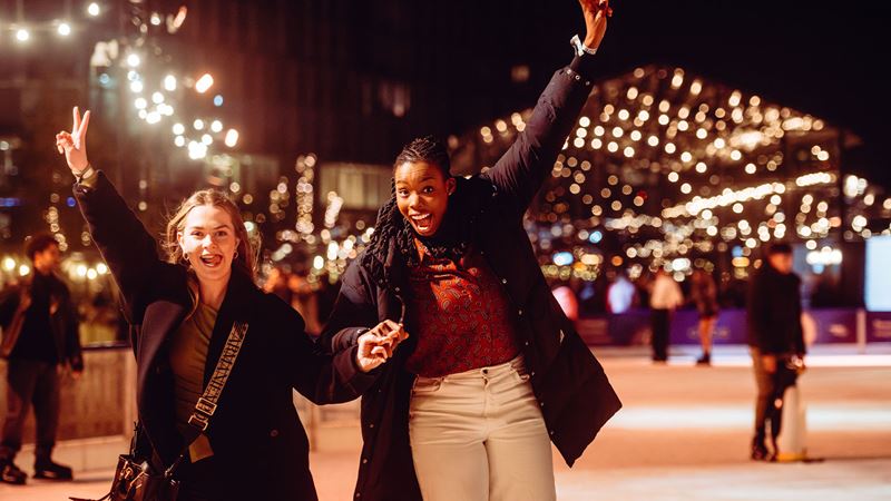 Two women are skating at the Glide ice rink in Battersea at night, enjoying their session and raising their hands in the air.