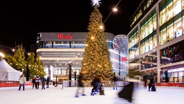 A few people are skating around a tall Christmas tree sitting in the middle of London West field's outdoor ice rink. 