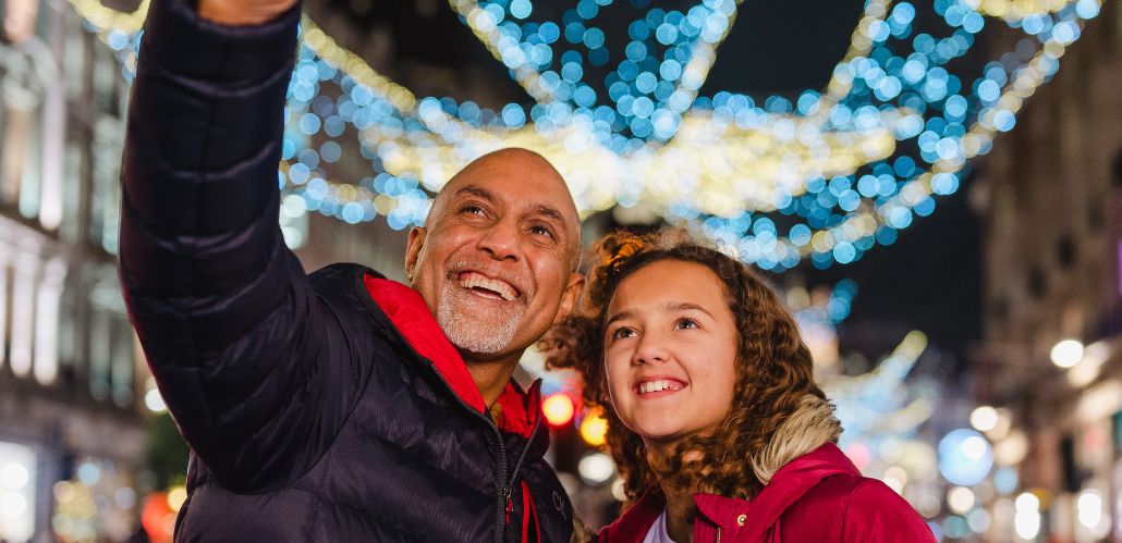 a man and young girl smiling up at a phone taking a selfie, with Christmas lights glowing in the background