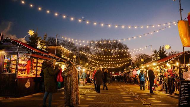 Winter Wonderland Christmas market. Image courtesy of Winter Wonderland. People dressed in coats visiting Christmas market stalls either side of twinkly string lights at dusk.