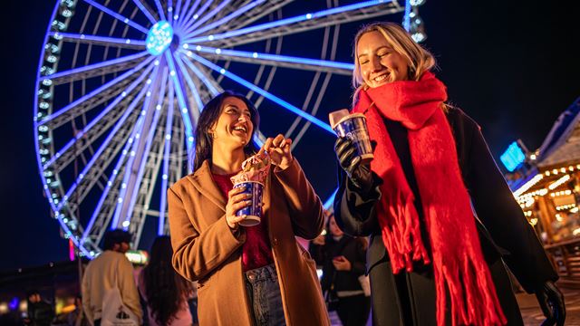 Two young women drinking hot chocolate in Winter Wonderland, with the Great Wheel in the background. 