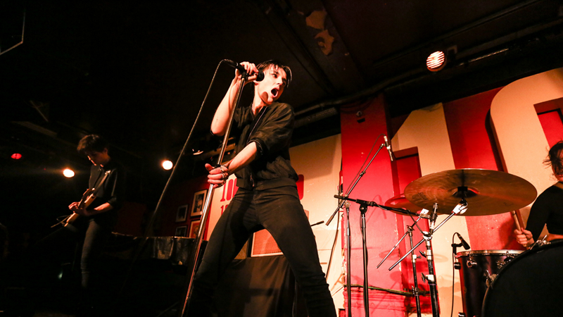 The Savages band performing live at The 100 Club. Credits: Neelam Khan Vela. Image courtesy of The 100 Club. A person holding a standing microphone on stage, singing, with red background.