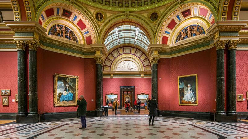 Wander the exhibition spaces of the National Gallery in London. Credit: National Gallery. Image courtesy of National Gallery. Two people looking at paintings in the red-painted halls of the National Gallery.