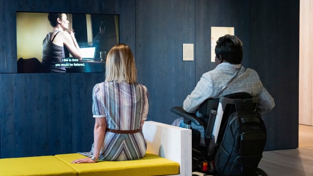 A woman in a striped dress sitting on a yellow bench, with a man on a wheelchair beside her. They are both watch a movie at an exhibition.