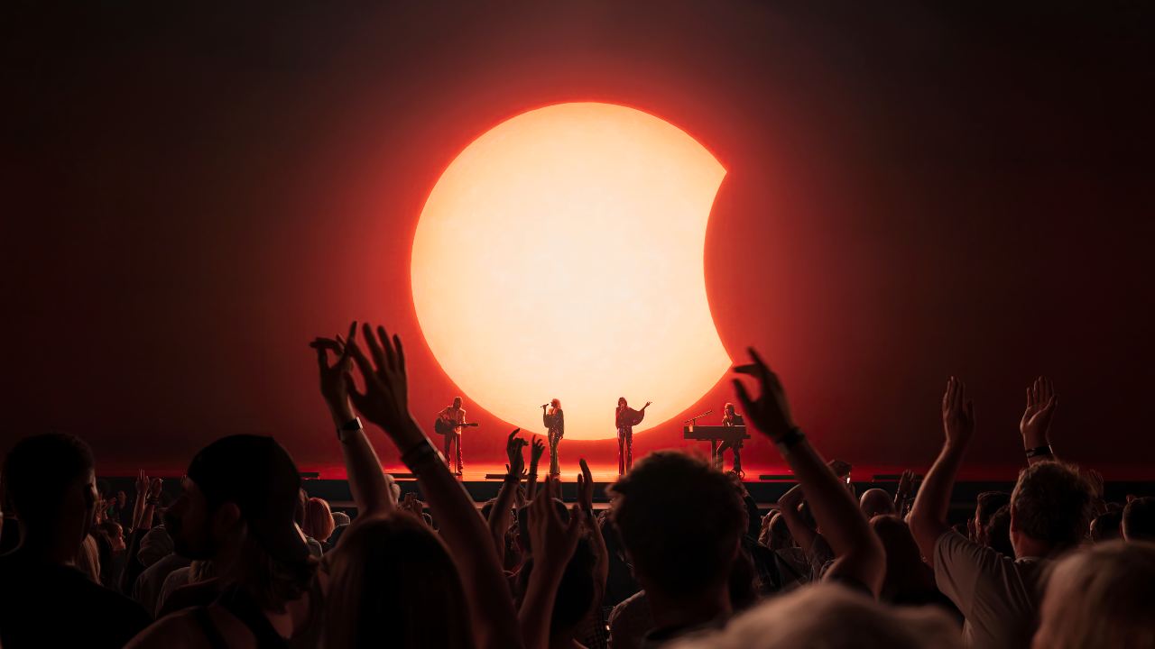 ABBA Voyage at London's ABBA Arena in Stratford. Image courtesy of ABBA Voyage/Johan Persson. ABBA Voyage performers on stage with red moon backdrop and live crowd with their arms up in the air at London's ABBA Arena.