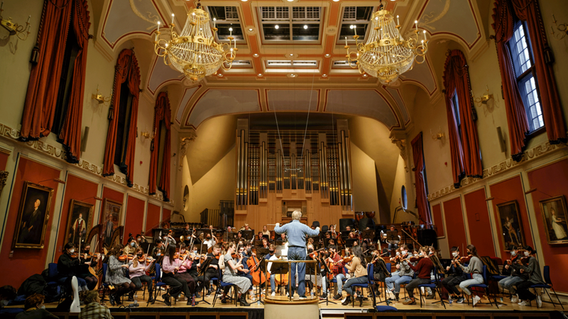 Sir Mark Elder conducts the Academy Symphony Orchestra. Credit: Craig Fuller. Image courtesy of Royal Academy of Music. Orchestra on stage being conducted with warm lights overhead