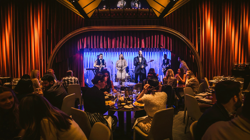 Tap your feet to the upbeat sounds of jazz and live singers at Quaglino's. Credit: D&D London. Image courtesy of D&D London. Band are illuminated with blue lights in front of tables of crowds