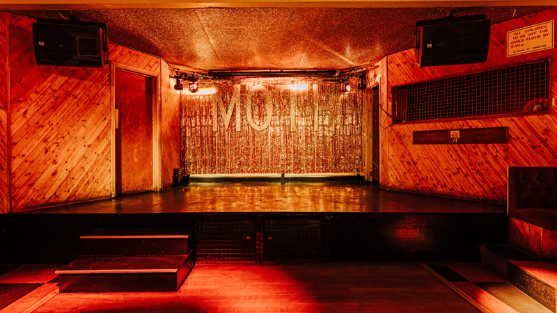 Join the crowds that flock to MOTH Club for rock nights in east London. Credit: Luke Dyson. Image courtesy of MOTH Club. Red lights illuminate the stage at MOTH Club with a glitter wall spelling out "MOTH"