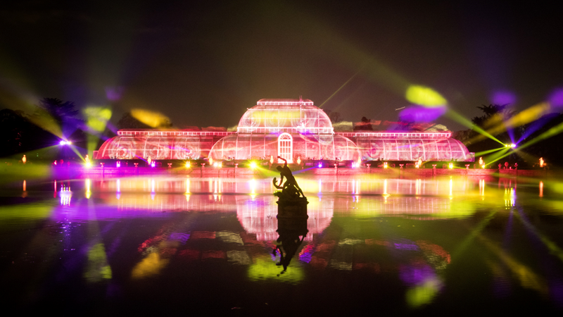 Wrap up and head to Christmas at Kew in west London to see colourful installations light up the iconic site. Credit: RBG Kew. Image courtesy of RBG Kew. Pink, orange and yellow lights are projected onto the greenhouse at Kew Gardens with reflections on the water with a statue in the middle