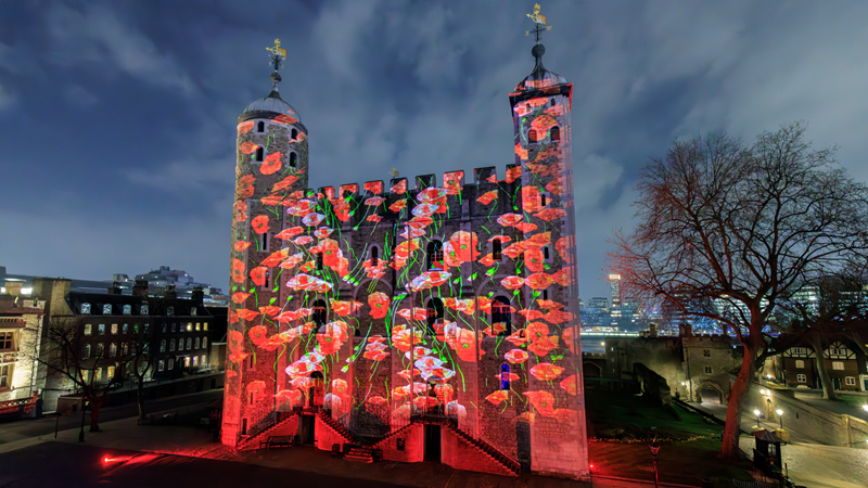 See Poppy Fields at the Tower this November. Image courtesy of Luxmuralis / Historic Royal Palaces. The Tower of London illuminated at night with red light show featuring poppies and shadows of soldiers to commemorate Remembrance in London.