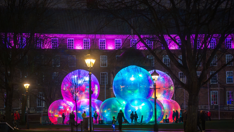 Step into a dreamlike world at Westfield London's Here We Glow, featuring light installations from five different artists. Credit: Andre Pattenden. Image courtesy of Westfield London. A collection of large transparent bubble sculptures glowing shades of blue, pink and purple at night with outlines of people and lamposts in the foreground