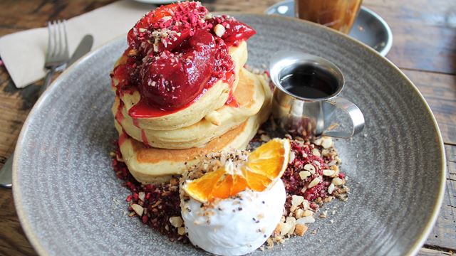 A stack of pancakes on a grey plate served with a silver pot of syrup and an orange segment on top of cream. 