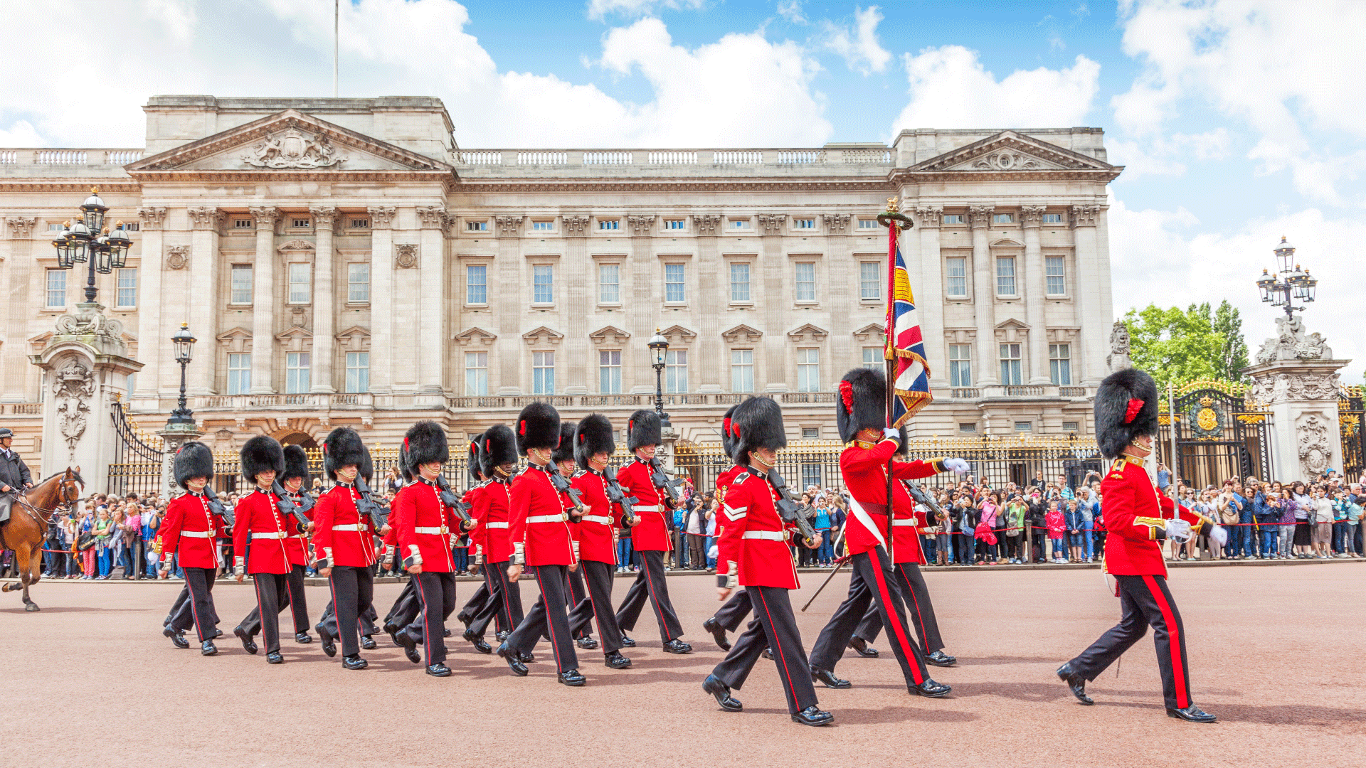 Watch the iconic Changing the Guard parade at Buckingham Palace. Credit: Shutterstock. Image courtesy of Shutterstock. Guards in traditional red tunics and bearskin hats form a line in front of Buckingham Palace holding a Union Jack flag