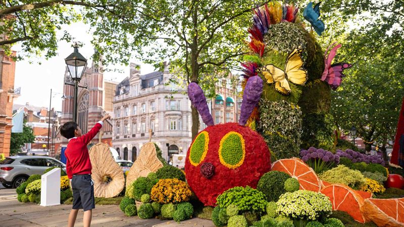 a young school boy wearing a red jumper and knee-length grey shorts, points up to a large floral installation of a Caterpillar, made out of brightly coloured flowers and plants